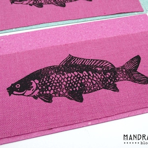 May include: Close-up of pink fabric with a black fish print. The fish is detailed with scales and fins. The fabric is on a light blue background. The text "MANDRAGORA block prints" is in the bottom right corner.
