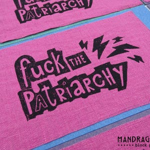 Könnte beinhalten: Rosa Stoffflicken mit dem Schriftzug "fuck the patriarchy" in schwarzen Blockbuchstaben und Blitzgrafiken. Die Flicken sind auf einer blauen und grünen Oberfläche angeordnet. Der Name des Künstlers, Mandragora, ist unten aufgedruckt.
