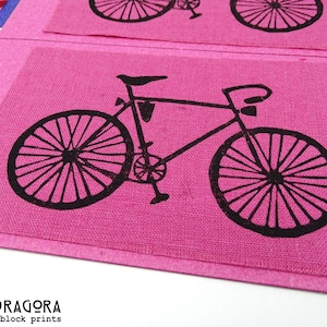 Pode incluir: Close-up de quadrados de tecido rosa com uma estampa de bicicleta preta. A bicicleta é um desenho simples com rodas detalhadas. Os quadrados estão dispostos em um padrão de grade. O texto "MANDRAGORA block prints" está no canto inferior esquerdo.