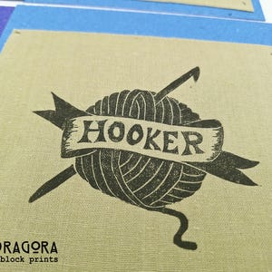 May include: A black and white block print on tan fabric features a yarn ball with two crochet hooks and a banner that reads "HOOKER". The print is on a square of fabric with a blue border. The bottom left corner has the text "MANDRAGORA block prints".