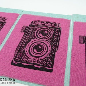May include: Pink fabric squares with a black illustration of a vintage camera. The camera design features two circular lenses and the text "Lubitel-2". The fabric is set against a light blue background.