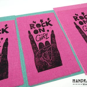 Pode incluir: Impressões em blocos de tecido rosa com o texto "ROCK ON, GIRL" em preto e um gesto de mão. As impressões são dispostas sobre um fundo turquesa. O canto inferior direito tem o texto "MANDRAGORA block prints".