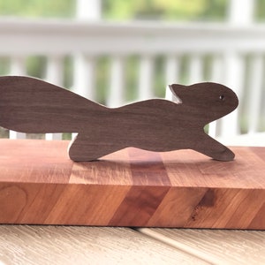 May include: A wooden squirrel figurine in a dark brown color, standing on a wooden cutting board with a chevron pattern.