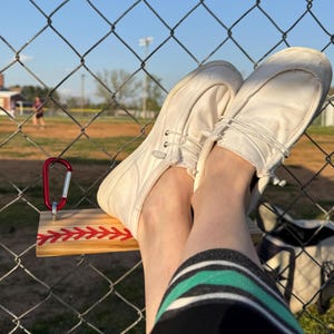 May include: A pair of white canvas shoes with laces are resting on a wooden plank with red stitching. The plank is attached to a chain link fence.