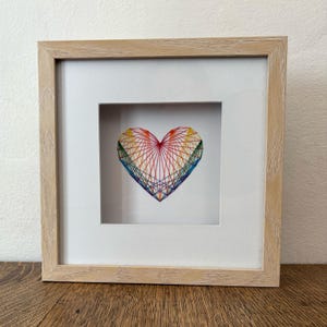 May include: A framed artwork featuring a heart design. The heart is crafted with colorful threads in a rainbow pattern, set against a white background. The frame is made of light-colored wood, adding a natural touch to the piece.