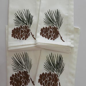 May include: A stack of four white cloth napkins. Each napkin features a brown pine cone and green pine needles design. The napkins are folded and stacked, showcasing the repeating nature-themed pattern.
