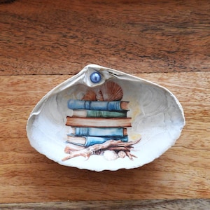 May include: A decorative seashell trinket dish with a painted design of a stack of books, seashells, and driftwood. The dish is white with a blue pearl accent. The background is a wooden surface.