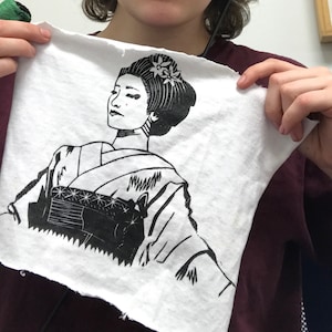May include: A black and white print of a woman in a traditional Japanese kimono. The print is on a white fabric with a slightly rough texture.