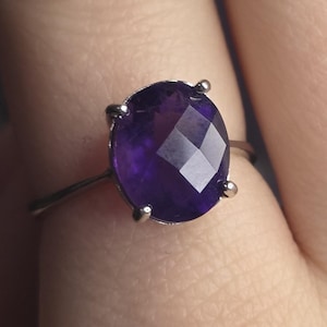 Puede incluir: Un anillo de plata con una gran gema de amatista facetada de corte ovalado. La piedra de color p&uacute;rpura oscuro est&aacute; sujeta por cuatro garras. El anillo se muestra en el dedo de una persona.