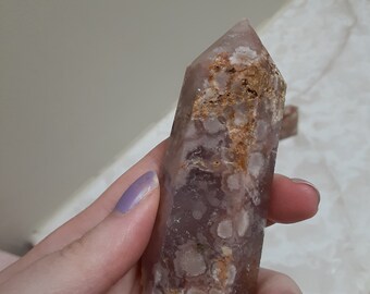 May include: A pink and brown agate crystal point with white and orange inclusions. The crystal is polished and has a smooth surface.