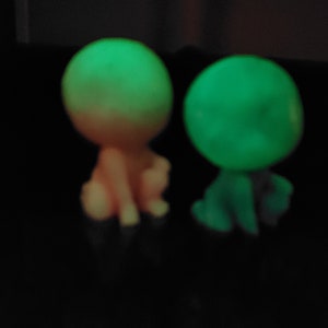 May include: Two small glow-in-the-dark figurines, one is pink and the other is green. They are both sitting down with their heads tilted.