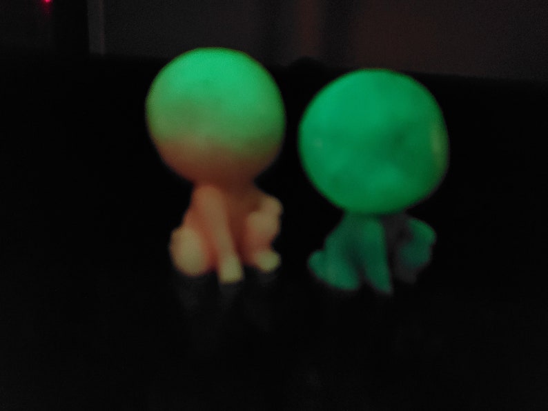 May include: Two small glow-in-the-dark figurines, one is pink and the other is green. They are both sitting down with their heads tilted.