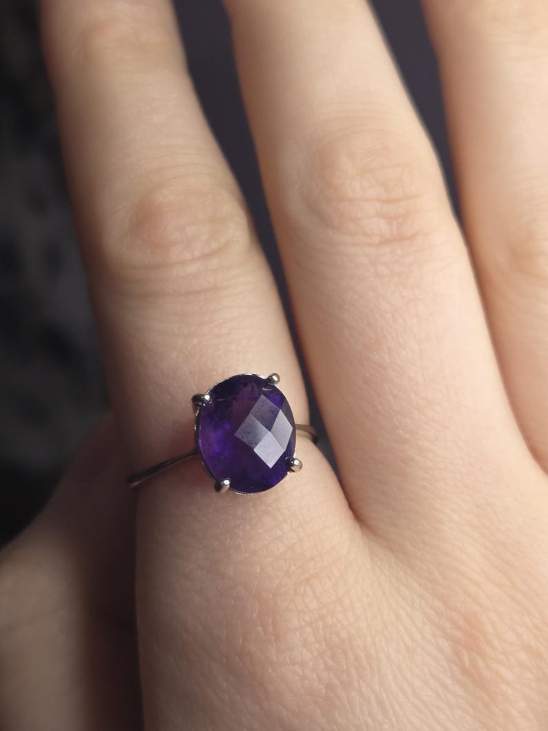 Puede incluir: Un anillo de plata con una gran gema de amatista facetada de corte ovalado. La piedra de color p&uacute;rpura oscuro est&aacute; sujeta por cuatro garras. El anillo se muestra en el dedo de una persona.