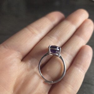 Puede incluir: Un anillo de plata con una banda ajustable, con una gran piedra preciosa de amatista p&uacute;rpura oscura de corte cuadrado. El anillo se sostiene en una mano, mostrando el color de la piedra preciosa y el dise&ntilde;o del anillo. Es una joya.