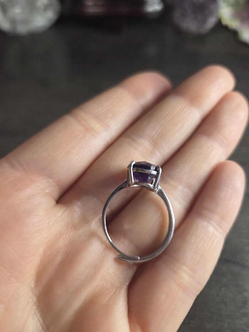 Puede incluir: Un anillo de plata con una banda ajustable, con una gran piedra preciosa de amatista p&uacute;rpura oscura de corte cuadrado. El anillo se sostiene en una mano, mostrando el color de la piedra preciosa y el dise&ntilde;o del anillo. Es una joya.