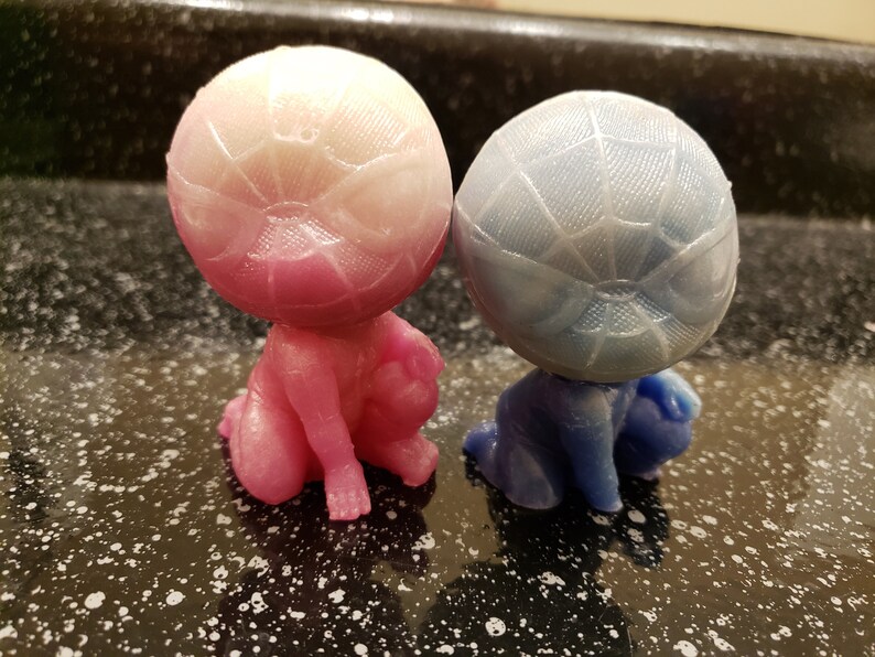 May include: Two small figurines, one pink and one blue, with a web-like pattern on their heads. They are sitting on a black surface.