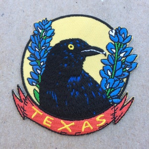 May include: Embroidered patch featuring a black bird with blue accents, surrounded by bluebonnets and a yellow circle. A red banner with the word "TEXAS" in yellow arcs across the bottom. The patch is likely for clothing or accessories.