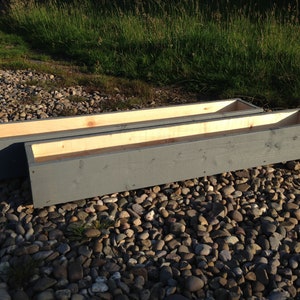 May include: Two long, rectangular wooden planters painted a dark grey colour. The planters are sitting on a bed of small, grey stones.
