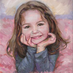 May include: A young girl with long brown hair smiles while resting her chin on her hands. She is wearing a blue long-sleeved shirt and is lying on a pink fluffy surface.
