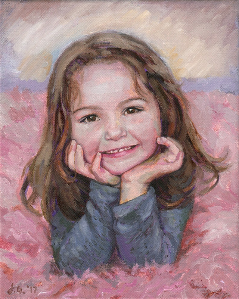 May include: A young girl with long brown hair smiles while resting her chin on her hands. She is wearing a blue long-sleeved shirt and is lying on a pink fluffy surface.