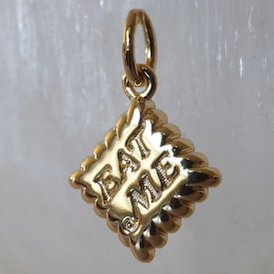 Puede incluir: Un colgante de metal dorado con forma de galleta cuadrada y bordes festoneados. El centro del colgante presenta el texto EAT ME en relieve con letras mayúsculas. Un pequeño anillo circular está sujeto en la parte superior para colgarlo.