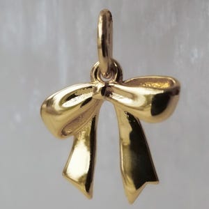 May include: A gold-colored bow pendant with a loop at the top for hanging. The bow has a detailed design with curved edges and two long, downward-pointing ribbon ends. The pendant is likely made of metal and has a shiny finish.