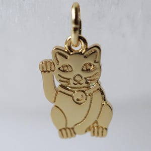 May include: A gold-colored charm in the shape of a Maneki-neko, or Japanese lucky cat, with its right paw raised. The charm has detailed features, including eyes, whiskers, and a collar with a bell. The charm is suspended from a small loop.