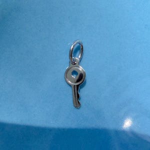 May include: A small, polished silver-toned metal charm shaped like a skeleton key. The charm features a circular head with a central heart-shaped cutout and a short, notched shaft. A circular jump ring is attached to the top of the key charm.