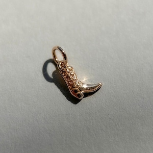 Könnte beinhalten: Ein kleiner, roségoldfarbener Metallanhänger in Form eines Cowboystiefels. Der Anhänger weist auf dem Schaft und dem Fuß detaillierte Prägungen auf und ist mit einem runden Biegering an der Oberseite zur Befestigung an einer Kette versehen.