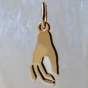 May include: A polished gold-toned metal pendant shaped like a hand with the thumb and index finger touching to form a circle. The pendant features a smooth, reflective surface and includes a small circular jump ring at the top for attachment.