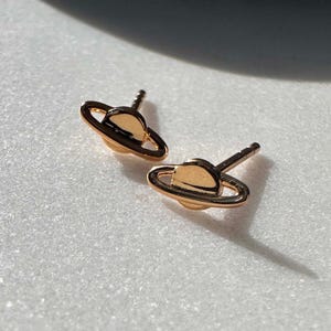 May include: A pair of gold-coloured stud earrings shaped like a planet with a ring. The earrings are small and delicate, with a shiny finish. They are set against a white background. Each earring is approximately 1 cm in diameter.