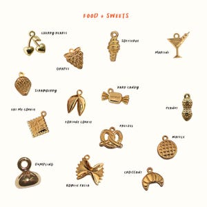 May include: A collection of gold-toned metal charms labeled Food + Sweets, including shapes like cherry hearts, grapes, soft serve, martini, strawberry, fortune cookie, hard candy, peanut, eat me cookie, pretzel, waffle, dumpling, bowtie pasta, and croissant.