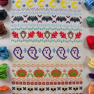 Peut inclure: Une broderie au point de croix blanche sur le th&egrave;me d'Halloween. Le motif pr&eacute;sente une vari&eacute;t&eacute; d'ic&ocirc;nes d'Halloween, notamment des fant&ocirc;mes, des citrouilles, des chauves-souris et des lunes. La broderie est entour&eacute;e de fils &agrave; broder color&eacute;s.