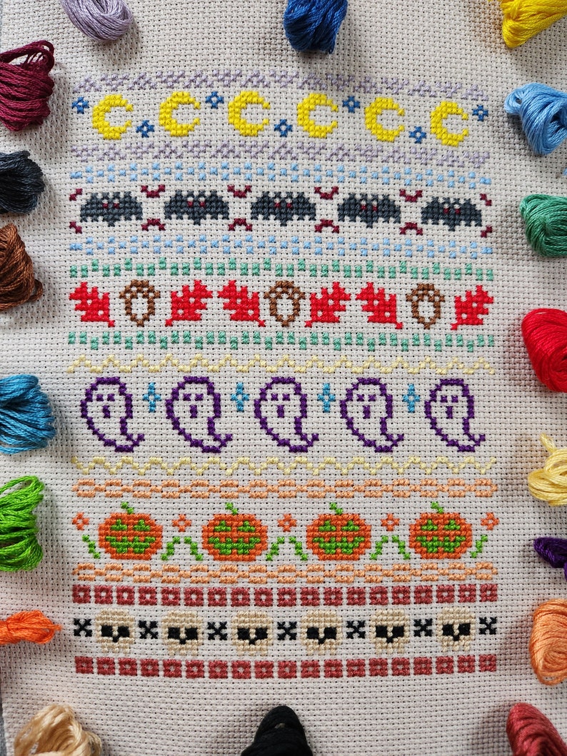 Peut inclure: Une broderie au point de croix blanche sur le th&egrave;me d'Halloween. Le motif pr&eacute;sente une vari&eacute;t&eacute; d'ic&ocirc;nes d'Halloween, notamment des fant&ocirc;mes, des citrouilles, des chauves-souris et des lunes. La broderie est entour&eacute;e de fils &agrave; broder color&eacute;s.