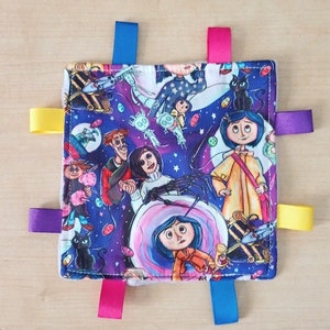 May include: A purple and blue baby blanket with a colorful print of characters from the movie "Coraline". The blanket has five colorful satin ribbons attached to the corners and sides.