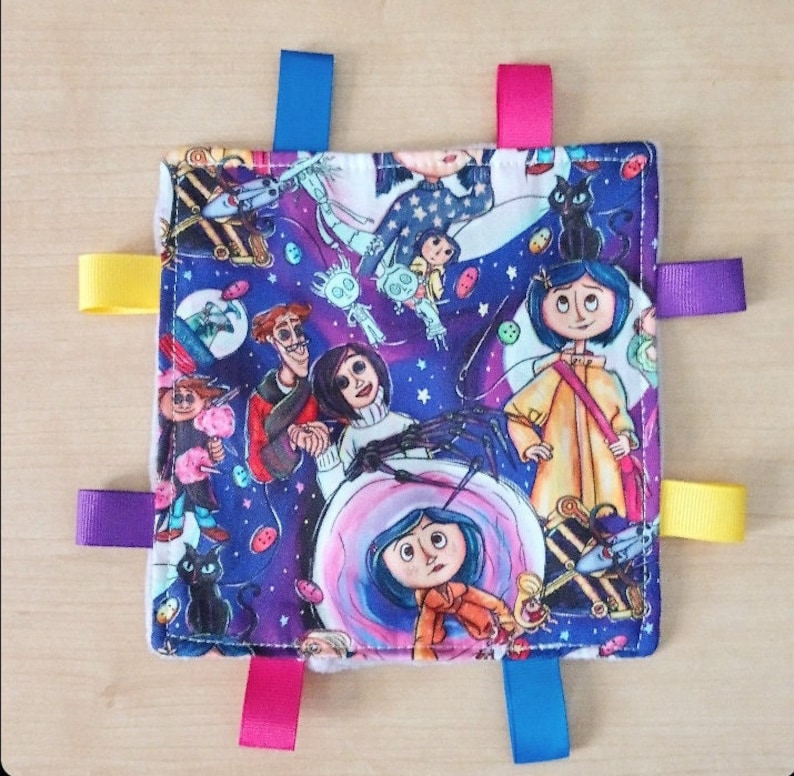 May include: A purple and blue baby blanket with a colorful print of characters from the movie "Coraline". The blanket has five colorful satin ribbons attached to the corners and sides.