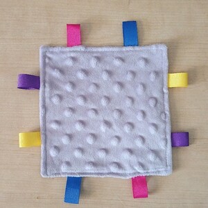 May include: A gray baby blanket with a textured surface and four colorful ribbon tags. The tags are purple, yellow, pink, and blue.