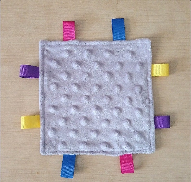 May include: A gray baby blanket with a textured surface and four colorful ribbon tags. The tags are purple, yellow, pink, and blue.