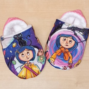 May include: A pair of baby slippers with a colourful print featuring a girl with a cat and a purple background. The slippers are made of soft fabric and have a pink lining.