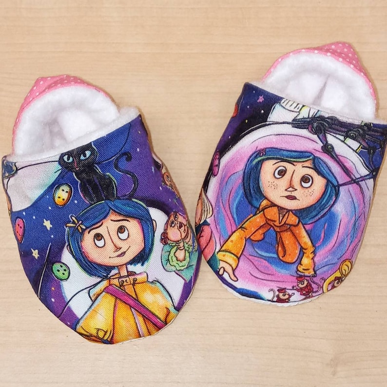 May include: A pair of baby slippers with a colourful print featuring a girl with a cat and a purple background. The slippers are made of soft fabric and have a pink lining.