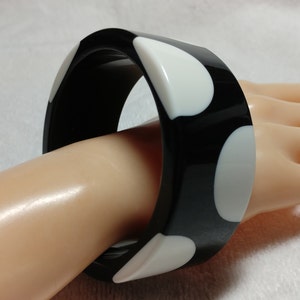 May include: A black and white bangle bracelet. The bracelet is a thick, square-shaped design with a glossy finish. White, rounded shapes are evenly spaced against the black background. The bracelet is shown on a person's hand.
