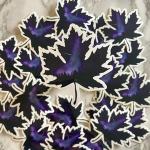 May include: A set of multiple stickers featuring a black maple leaf with a purple and blue galaxy inside. The stickers are perfect for adding a touch of nature and whimsy to laptops, water bottles, or journals.