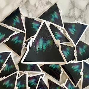 Peut inclure: Une pile d'autocollants en forme de triangle avec une silhouette de montagne noire et un motif d'aurore boréale coloré. Les autocollants sont imprimés sur du papier blanc avec une bordure blanche.