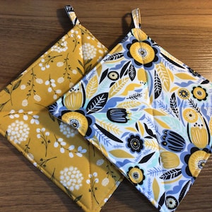 May include: Two quilted pot holders with floral patterns. One is yellow with white flowers and the other is white with blue, yellow, and black flowers.