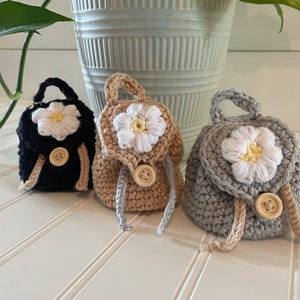 May include: Three crocheted mini backpacks with daisy flower accents and wooden button closures. The backpacks are black, beige, and gray.