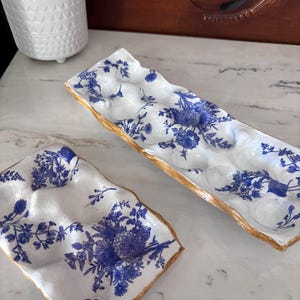 Pode incluir: Duas travessas de ovos de cerâmica branca com padrões florais azuis e acabamento dourado. Uma travessa é retangular, a outra quadrada. As travessas têm compartimentos moldados para ovos. As travessas estão sobre uma superfície de mármore.