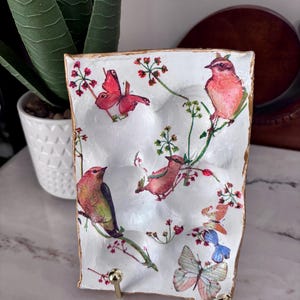 May include: A white, rectangular decorative tray with a gold rim, featuring a watercolor design of pink birds and butterflies on branches with red flowers. The tray is supported by a gold metal stand.