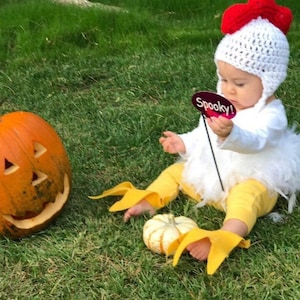 Könnte beinhalten: Ein Baby, das sich für Halloween als Huhn verkleidet hat, hält ein Schild mit der Aufschrift "Spooky!" Das Baby trägt eine weiße Häkelmütze mit rotem Kamm und gelben Hühnerfüßen. Das Baby sitzt auf grünem Gras neben einem geschnitzten Kürbis.