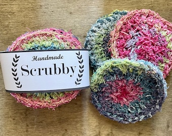 Op de afbeelding: Drie handgemaakte schuursponsen in verschillende kleuren, waaronder roze, groen en blauw. De sponzen zijn gemaakt van gerecyclede materialen en zijn perfect voor het schoonmaken van vaat, aanrechtbladen en andere oppervlakken. Het label luidt "Handmade Scrubby".