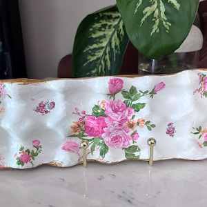 May include: A white, rectangular decorative tray with a gold-painted edge. The tray is adorned with pink roses and floral accents. Two gold-colored hooks are attached to the front. A decorative piece for the home.